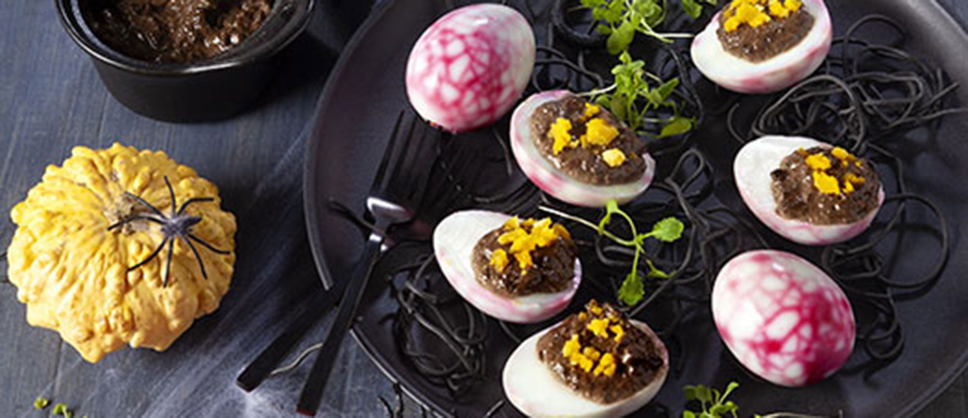 Demi-œufs durs aux blancs marbrés de rose, garnis de mayonnaise noire et de jaune émietté, sur un lit de spaghettis noirs, à côté d'une petite courge et d'une araignée en plastique